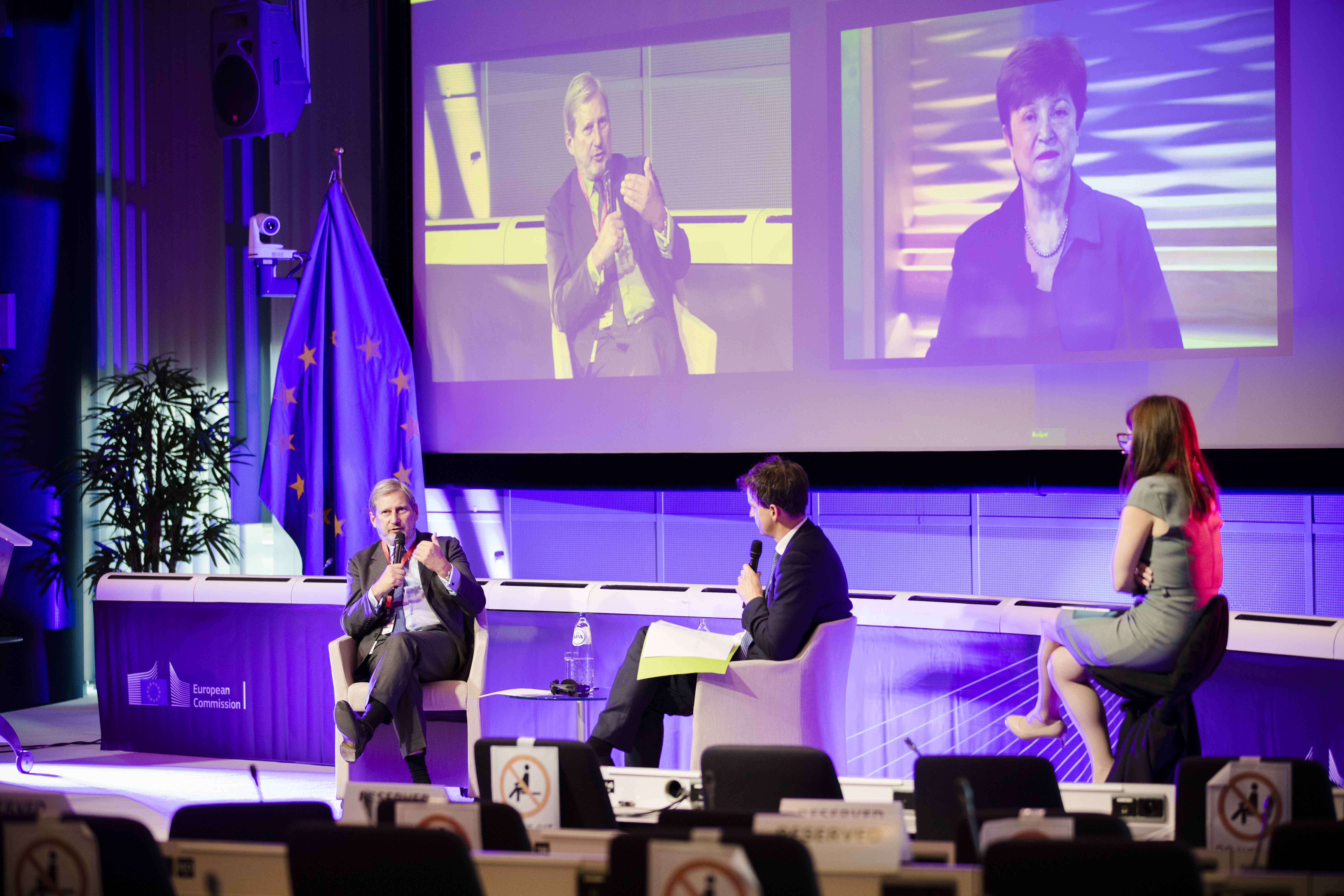 stage + screen, 3rd panel discussion, from left to right: Commissioner Johannes Hahn,  moderator Sam Fleming, (Brussels Bureau Chief, Financial Times), master of ceremony Beatriz Rios. On the screen, on the right side IMF Managing Director, Kristalina Geo