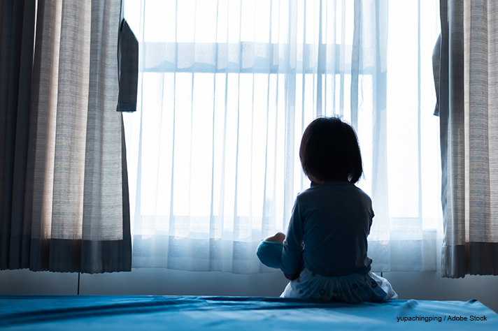 Małe dziecko siedzi na łóżku bawiąc się lalką w ciemnym pokoju z lekką kurtyną