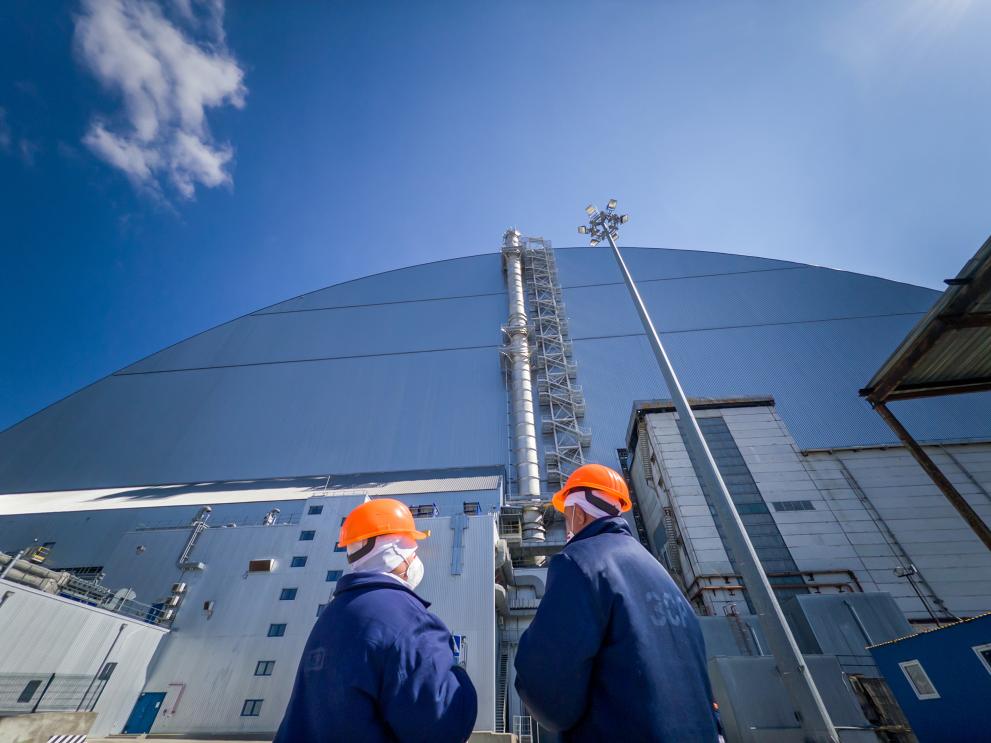 Two people in protective gear stand in front of the New Safe Confinement at Chernobyl 