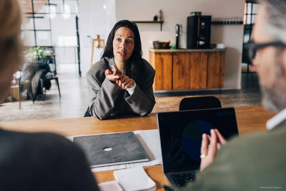 A woman gestures while speaking with two colleagues across an office table during a job interview