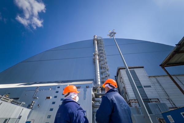 Two people in protective gear stand in front of the New Safe Confinement at Chernobyl 